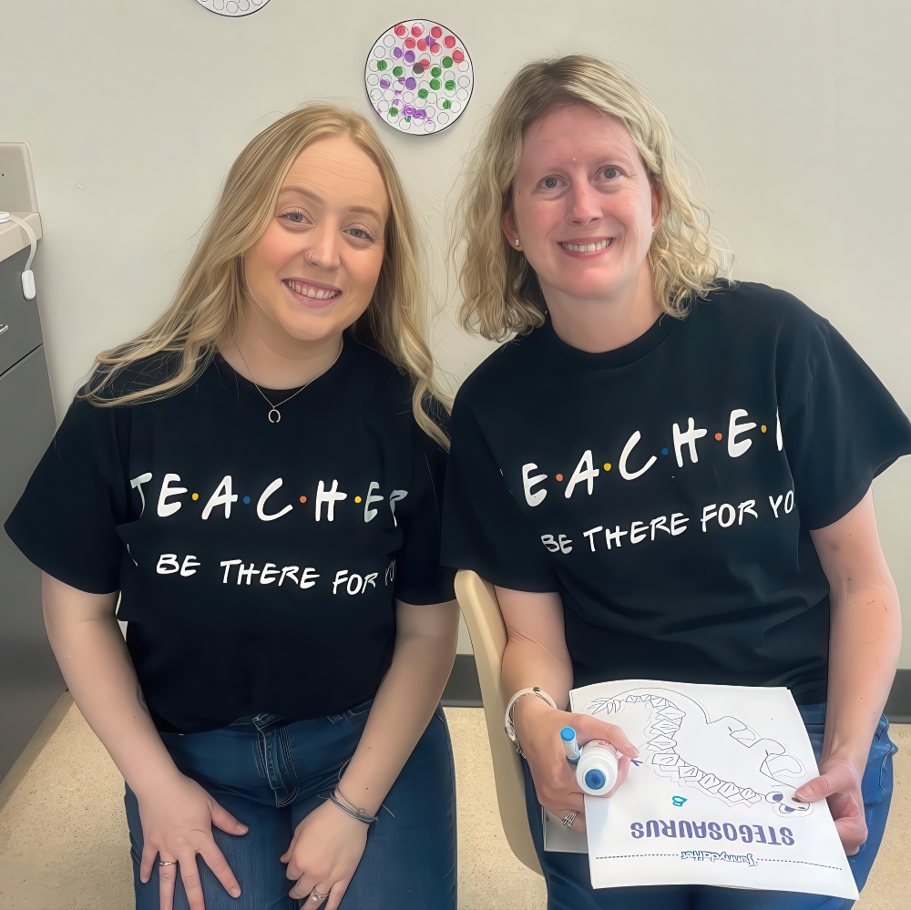 Two teachers smile in matching shirts that say “TEACHER.”