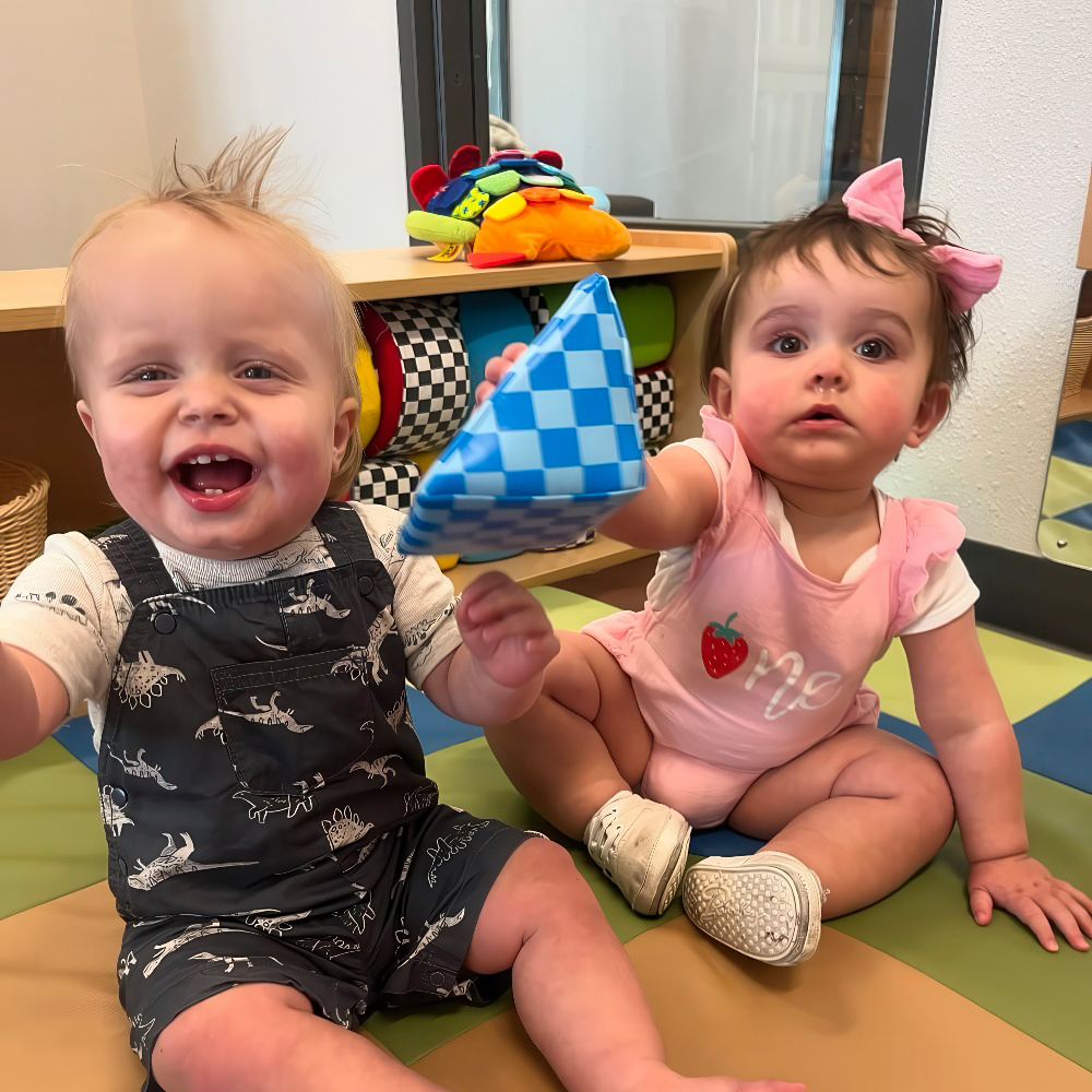 Two babies smile and play with blue patterned blocks, teacher close.