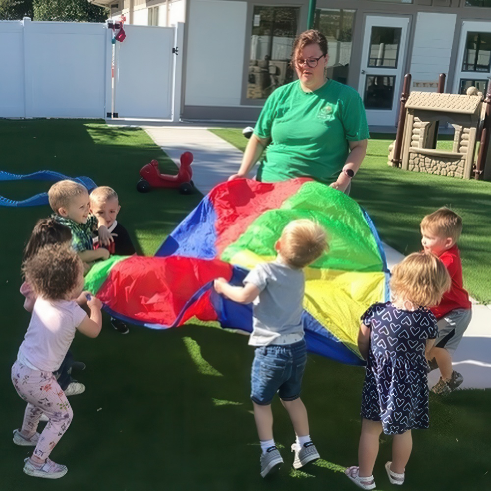 A teacher leading a group of children in a colorful parachute play activity outside.