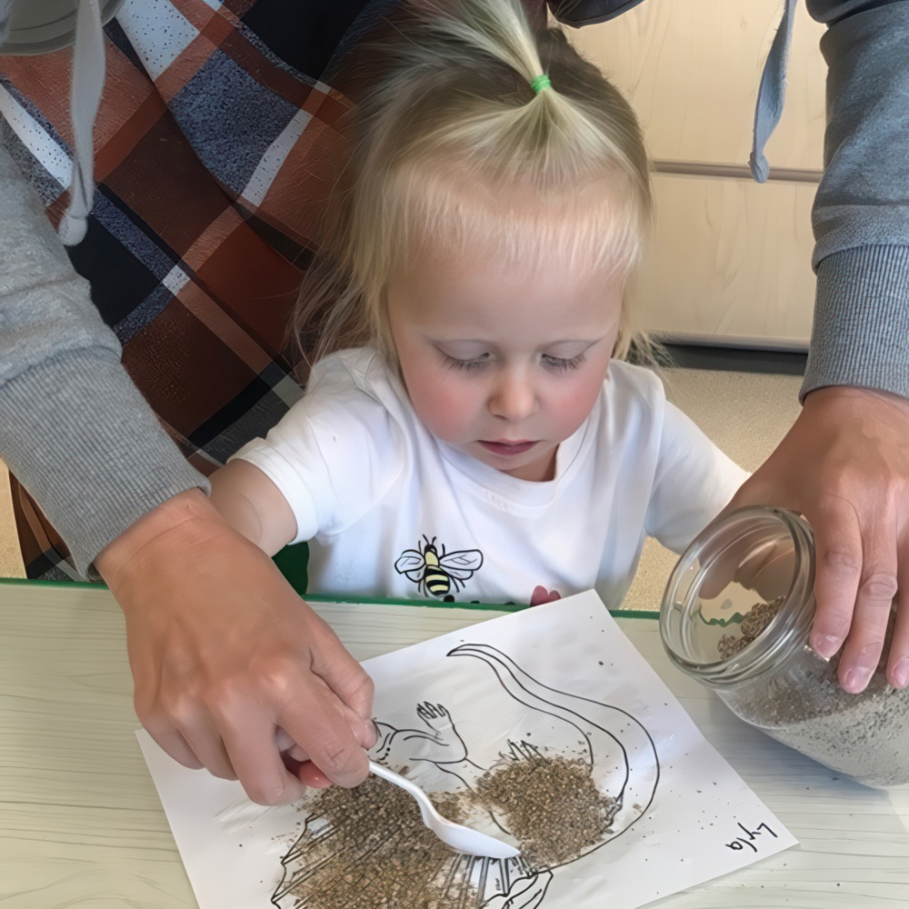 Teacher assists child sprinkling seeds onto a project sheet.